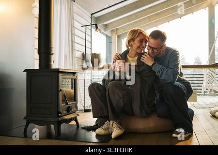 Caucasian middle aged woman sitting on floor holding mug, smiling while being embraced by Caucasian middle aged man, both relaxing together near wood burning stove in cozy cabin Stock Photo