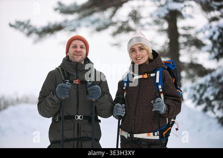 Middle aged Caucasian man and middle aged Caucasian woman standing outdoors holding trekking poles, wearing backpacks and winter hats, smiling while posing in snowy forest environment Stock Photo