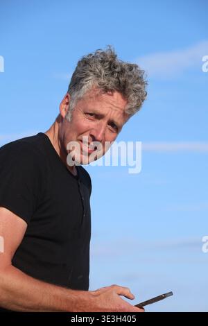 Portrait of a smiling middle-aged man holding a smartphone outdoors. Bright natural light, clear sky and relaxed atmosphere, lifestyle, communication Stock Photo