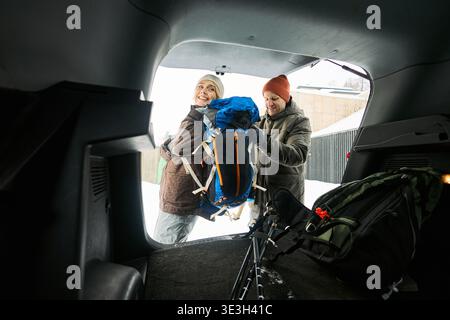 Middle aged Caucasian woman and middle aged Caucasian man loading hiking backpack into car trunk, standing outdoors in snowy environment, preparing for winter adventure or travel Stock Photo