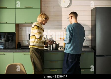 Caucasian middle aged woman and adult man preparing food together in modern kitchen, woman pouring ingredient into mixing bowl while man holding plate and standing nearby Stock Photo