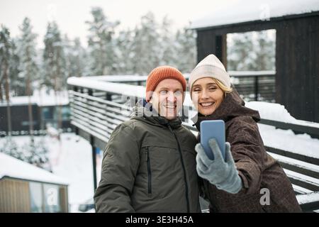 Middle aged Caucasian man and middle aged Caucasian woman smiling and taking selfie outdoors in snowy winter setting, both wearing warm hats and jackets, standing close together Stock Photo
