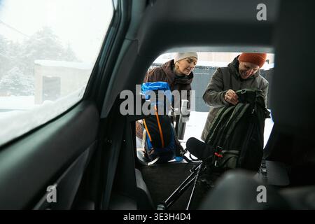 Middle aged Caucasian man and middle aged Caucasian woman packing backpacks into car trunk, preparing for winter outdoor activity, standing together in snowy driveway, smiling and interacting Stock Photo