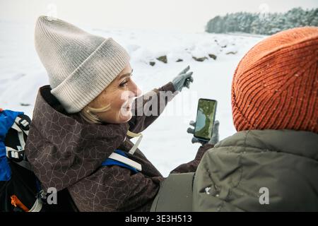 Caucasian adult woman smiling while holding smartphone and pointing toward snowy landscape, standing outdoors with another during winter hike, wearing winter clothing Stock Photo