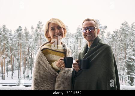 Caucasian woman and man standing outdoors wrapped in blankets smiling and holding black mugs with snowy forest background visible behind them Stock Photo