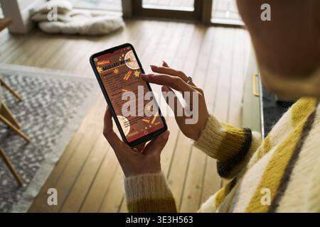 Caucasian middle aged woman using smartphone, holding device in left hand while scrolling with right index finger, reading mulled wine recipe displayed on screen indoors Stock Photo