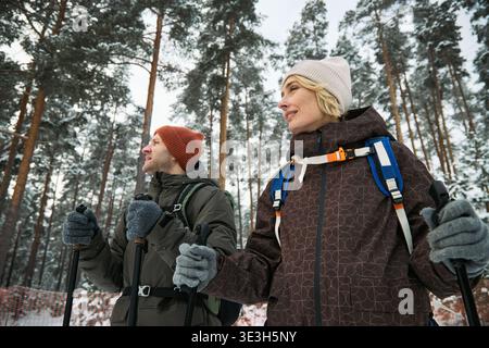Middle aged Caucasian man and middle aged Caucasian woman hiking with trekking poles in snowy forest, both wearing backpacks and winter hats, looking into distance among tall pine trees Stock Photo