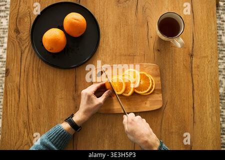 Man slicing fresh orange on wooden cutting board with knife, placing orange slices next to whole oranges on black plate, cup of tea standing on table nearby Stock Photo