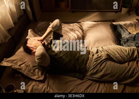 Adult Man Lying on Bed Covering Face with Hands in Bedroom Stock Photo