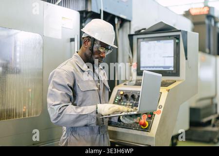 Professional African black worker, Engineer male working control operate CNC metal milling machine in industry factory with laptop computer. Stock Photo