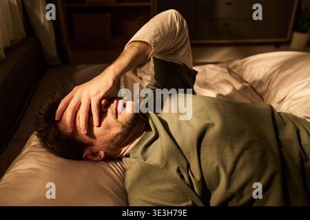 Young Man Lying in Bed Covering Face with Hand in Morning Stock Photo