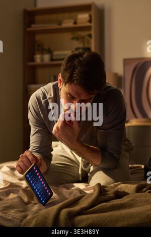 Young Man Sitting on Bed Checking Smartphone Notifications at Night Stock Photo