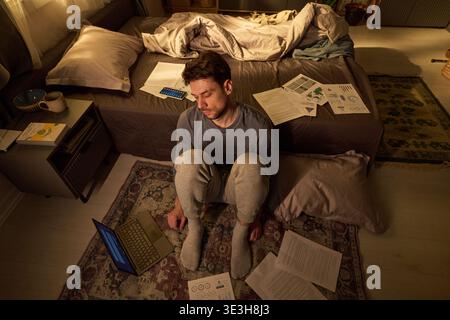 Young Man Sitting on Floor Working Late Night with Laptop and Papers Stock Photo