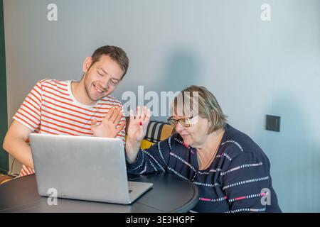 Adult son and elderly mother sitting at table using laptop and waving during video call with family. Remote communication, family connection and Stock Photo