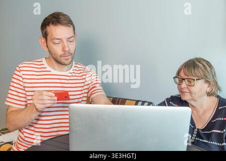 Adult son teaching his elderly mother how to make online payment using laptop and credit card. Digital banking, financial technology learning Stock Photo