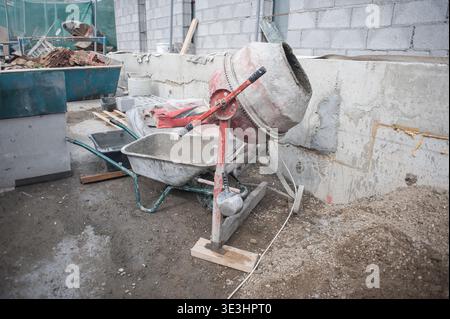 Cement mixer rests next to a freshly laid foundation on a construction site, surrounded by gravel, tools, and piles of materials. Workers prepare for Stock Photo