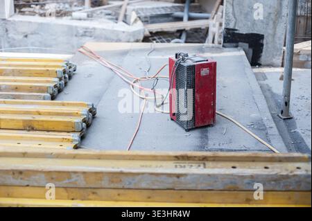 Construction tools and materials are scattered across a bustling site. A compact heater stands on a concrete surface, providing warmth as workers buil Stock Photo