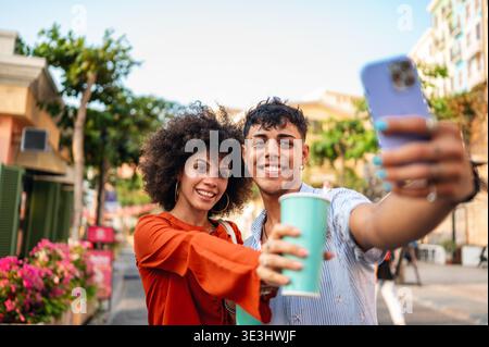 Diverse young couple happily taking a selfie on a vibrant street, capturing their joyful travel Stock Photo