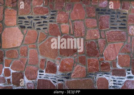 Red Stone Masonry Wall Texture Pattern Background. Full frame view of a rustic red stone wall with mortar joints and dark inserts, ideal as Stock Photo