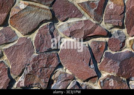 Rustic Red Stone Wall Texture Background. Full frame close-up of an irregular red stone wall with mortar joints, rough natural texture, and rustic Stock Photo