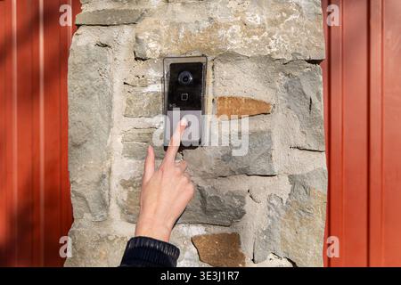 Finger Pressing Video Doorbell on Stone Gate. Close-up of a hand pressing a smart video doorbell on a stone wall, showing home security, access Stock Photo