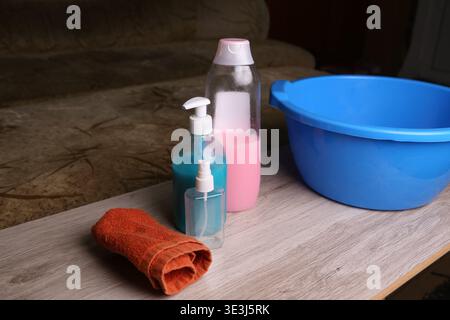 Cleaning supplies are placed on a table beside a blue bowl. There are bottles with different colored liquids and an orange towel ready for use in a cleaning task Stock Photo