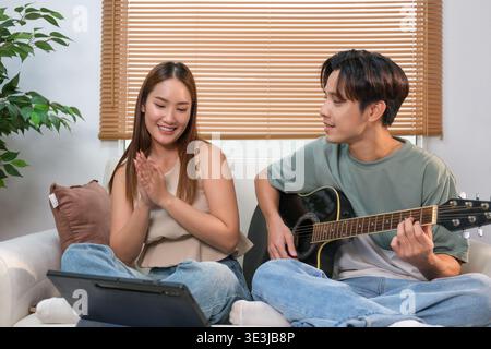 A young Asian couple spends their free time playing guitar and singing at home. Stock Photo
