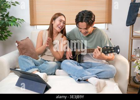 A young Asian couple spends their free time playing guitar and singing at home. Stock Photo