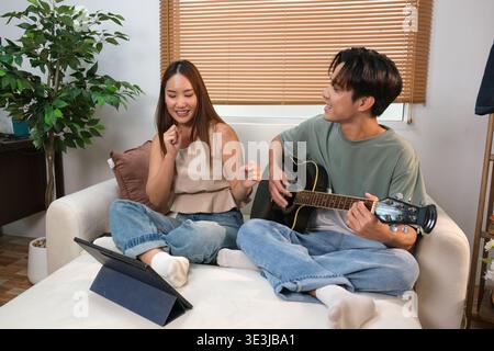 A young Asian couple spends their free time playing guitar and singing at home. Stock Photo