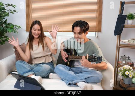 A young Asian couple spends their free time playing guitar and singing at home. Stock Photo