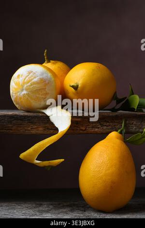 Vertical Lemon Still Life. Four lemons one partially peeled with its rind hanging below the rustic wood table. Side light against a mottled warm brown Stock Photo
