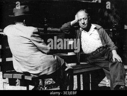 In this 1940s black and white photograph by Angelo Rizzuto, two men are seated on a park bench, capturing a moment of everyday life in New York. One man faces away from the camera, wearing a hat and light-colored jacket, while the other rests his head on his hand, appearing contemplative or tired. Stock Photo