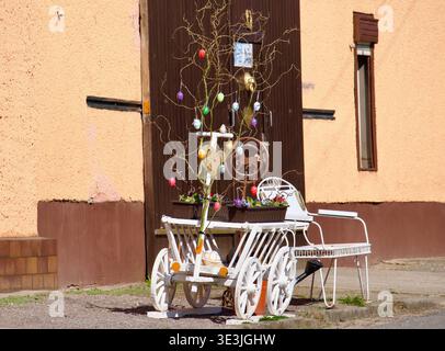 A rustic outdoor Easter display featuring a white vintage wooden decorative cart and a "Osterstrauch" (Easter tree) adorned with colorful hanging eggs. Set against a traditional house wall in a village. German Easter Decoration with White Wooden Cart and Decorated Tree. High quality photo Stock Photo
