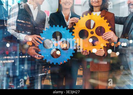 Team members hold large blue and orange gears in a modern office setting during a collaborative project meeting Stock Photo