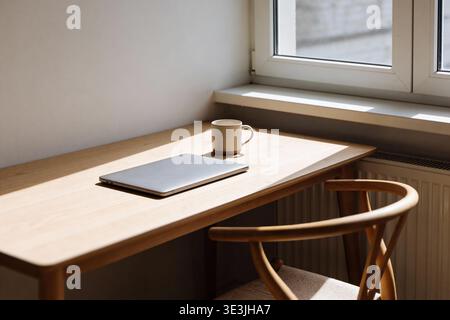 A clean and uncluttered workspace featuring a closed laptop and a ceramic mug on a light wood desk, illuminated by soft window light Stock Photo