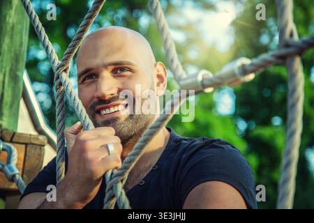A portrait of a smiling, bearded man in a black polo shirt in a natural setting. He looks at the camera from behind a mesh of thick ropes. The backgro Stock Photo