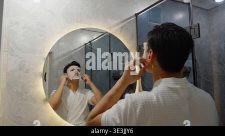 Young man applying face cream while looking in bathroom mirror Stock Photo