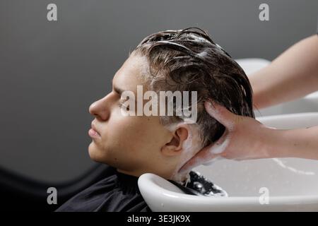 Hairdresser washes a boy hair with shampoo at a salon sink. Side view of scalp massage, hair care routine, hygiene, and grooming service in barbershop Stock Photo