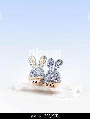 Five Easter eggs with funny faces and gray crocheted hats with bunny ears in a white egg stand on a gray background. Happy Easter concept. Greeting card. Stock Photo