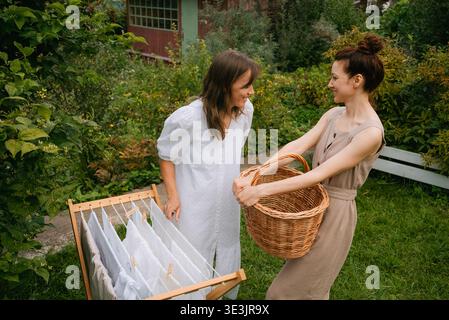 Two smiling women are hanging clean white laundry on a wooden clothesline in a green garden, one holding a wicker basket, enjoying a sunny summer day together Stock Photo