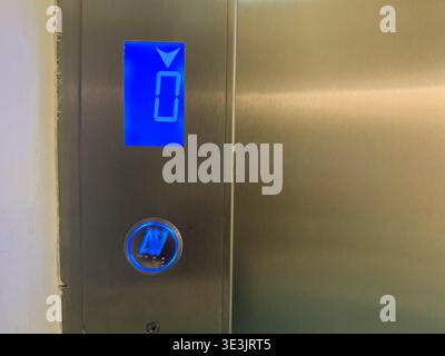 Stainless steel elevator panel with blue digital floor indicator showing zero beside illuminated call button. Vertical transportation access, building Stock Photo