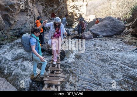 A group of hikers help each other across a narrow makeshift wooden bridge over a fast rocky mountain stream in a canyon, illustrating teamwork, advent Stock Photo