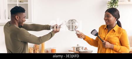 Positive african american couple having fun in kitchen, playfully fighting with kitchenware Stock Photo