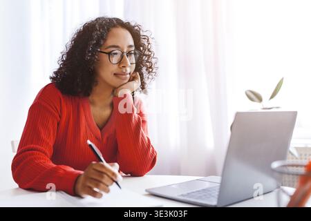 Woman Sitting at Table Working on Laptop While Taking Notes in a Bright Room During the Day Stock Photo