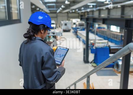 An Indian female engineer stands in a factory holding a tablet. She monitors machinery and operations in a manufacturing area. Stock Photo