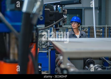 An Indian female engineer works in a factory wearing safety gear while operating machinery in a busy industrial environment. Stock Photo
