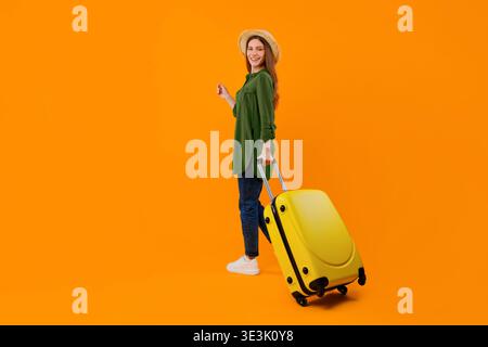 Woman With Suitcase Stands Against Bright Orange Background While Smiling Stock Photo