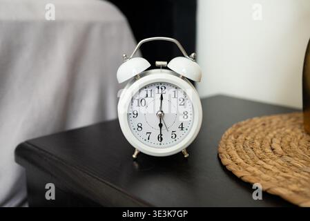 A classic white alarm clock with silver accents sits on a sleek black nightstand next to a woven mat and a white wall in the background, capturing a serene morning moment. Stock Photo