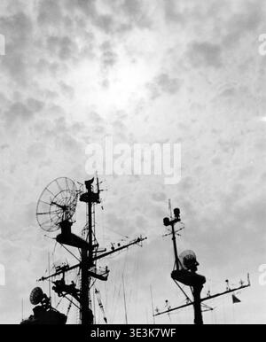 This vintage photograph, likely from the 1960s, captures a dramatic, low-angle silhouette of a ship's radar antenna and various electronic equipment against a cloudy sky. The intricate mast and dish antennas evoke a sense of advanced technology for its time, potentially from New York Harbor. Stock Photo