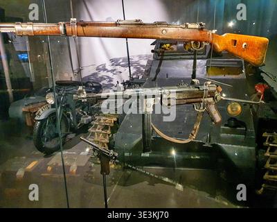 A museum display case featuring World War II military weapons and vehicles. Three firearms are suspended in the foreground: a wooden-stocked Stock Photo